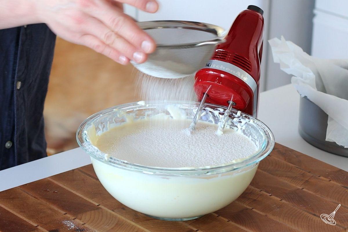 Someone sifting flour into a large bowl of cheesecake batter. 