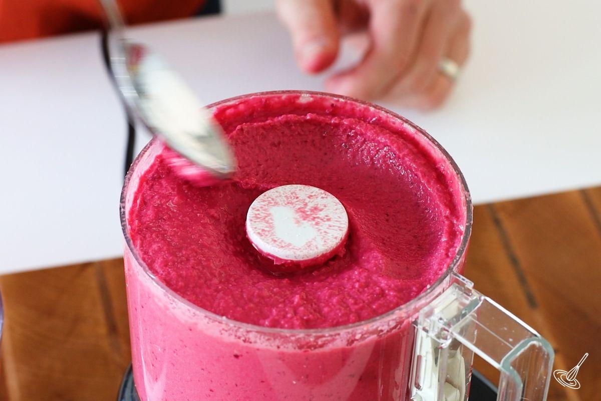 Someone running a spoon on top of a food processor of Lemon Beet Hummus.
