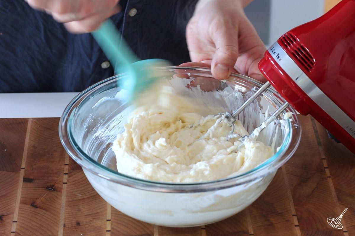 Someone using a spatula to scrape down a bowl of cream cheese. 