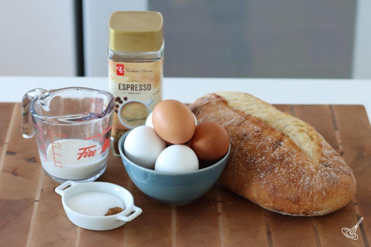 Ingredients on a cutting board, including a sourdough bread, eggs, milk, and espresso powder.