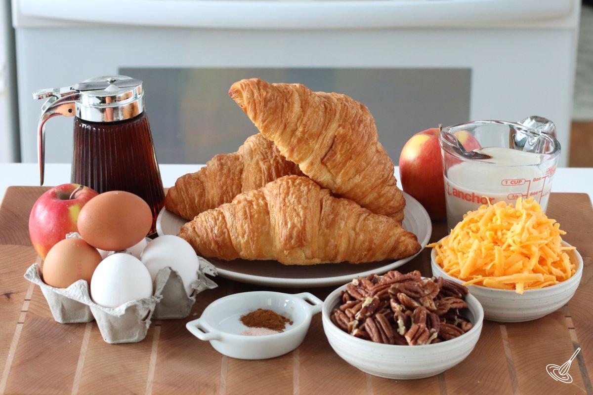 Ingredients on a cutting board, including croissant, grated cheese, eggs, and pecans.