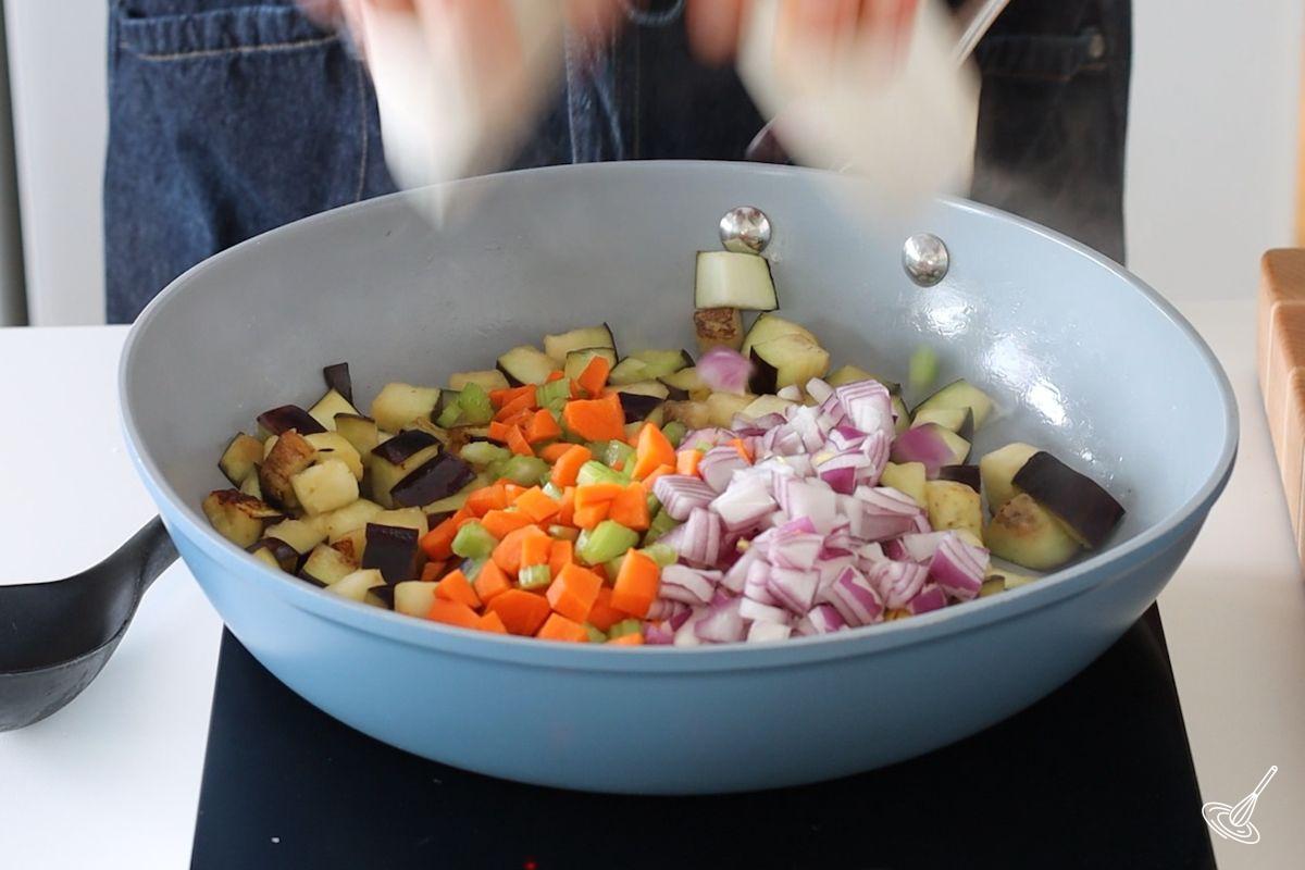 Someone adding chopped vegetables to a large pan of cooked aubergine cubes.