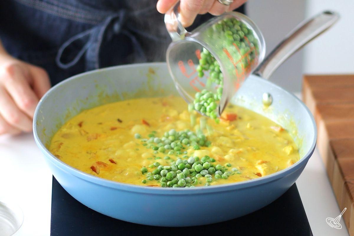 Someone adding frozen peas to a large pan of chicken curry sauce. 