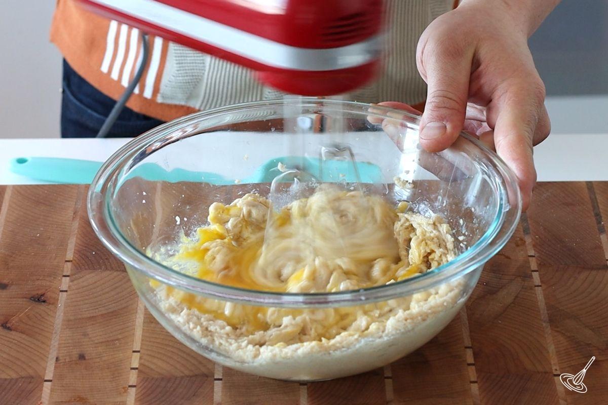 Someone using a hand blender to mix eggs into a butter and sugar mixture.