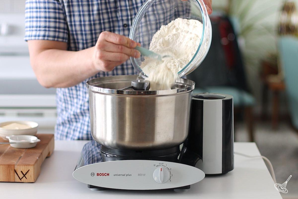 Someone adding flour into the bowl of a stand mixer.