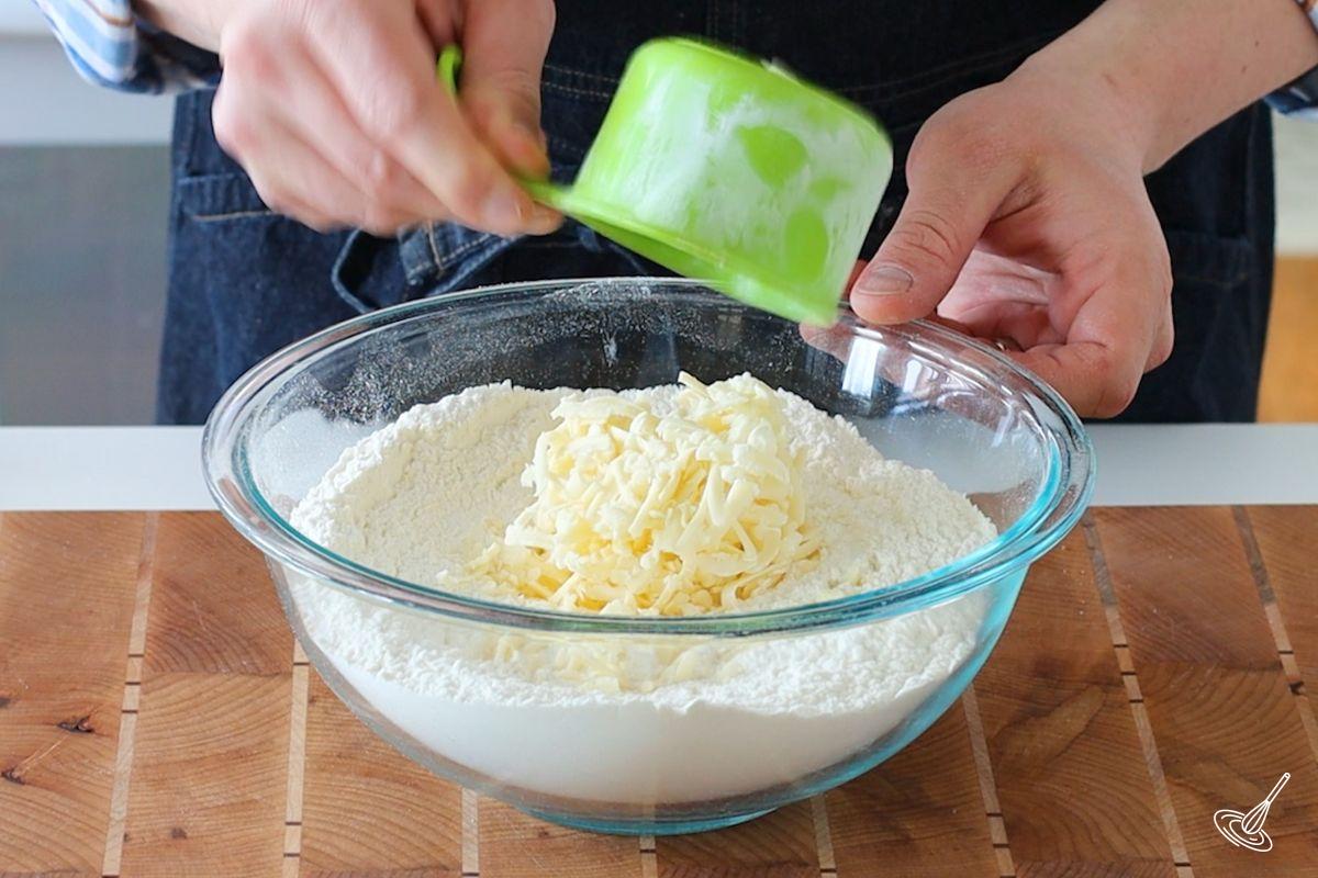 Someone adding grated cheese to a large bowl of flour. 