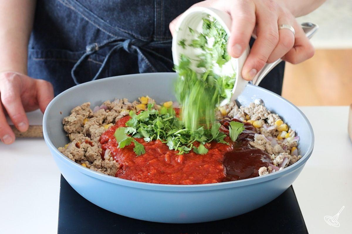 Someone adding fresh cilantro to a large pan of meat sauce.