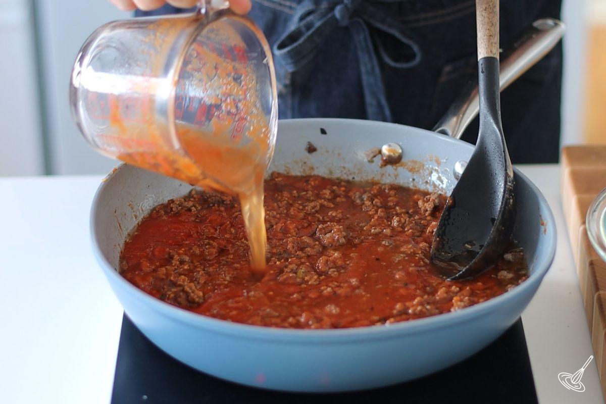 Someone adding water to a large pan of meaty pasta sauce. 