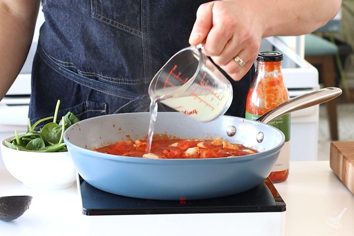 Someone pouring pasta water in a large skillet with tomato sauce.