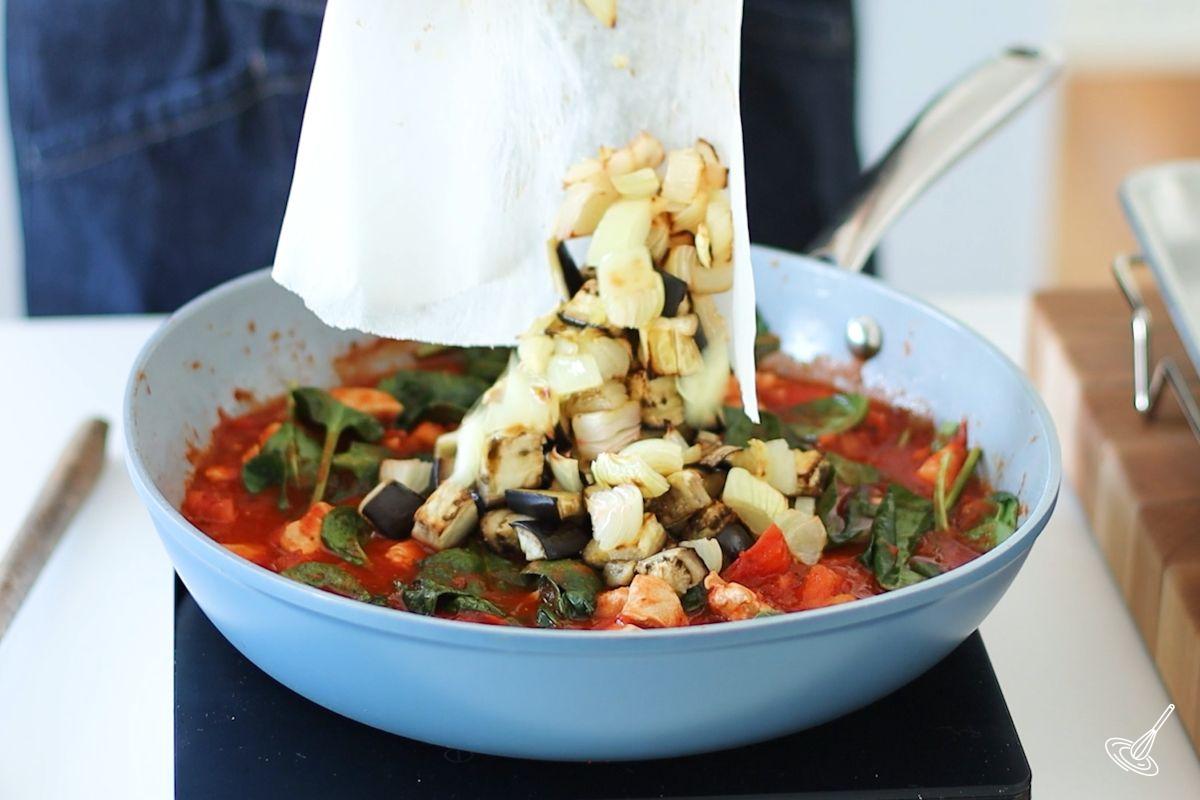Someone adding roasted vegetables to a skillet of pasta sauce.