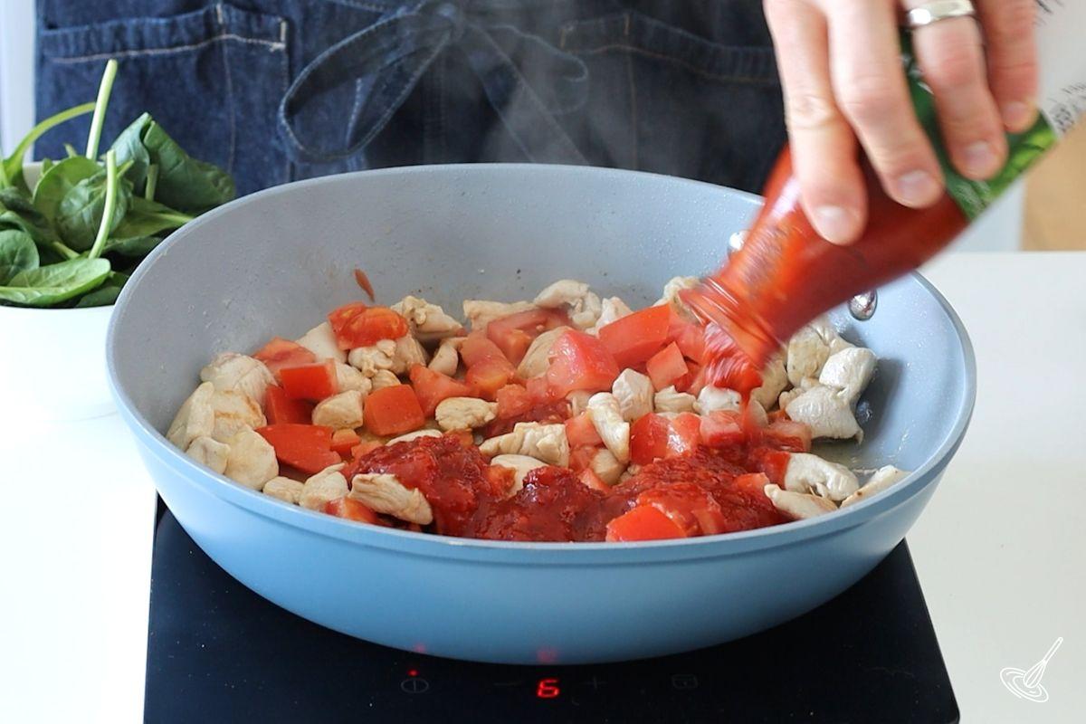 Someone pouring passata in a large skillet with tomatoes and chicken.