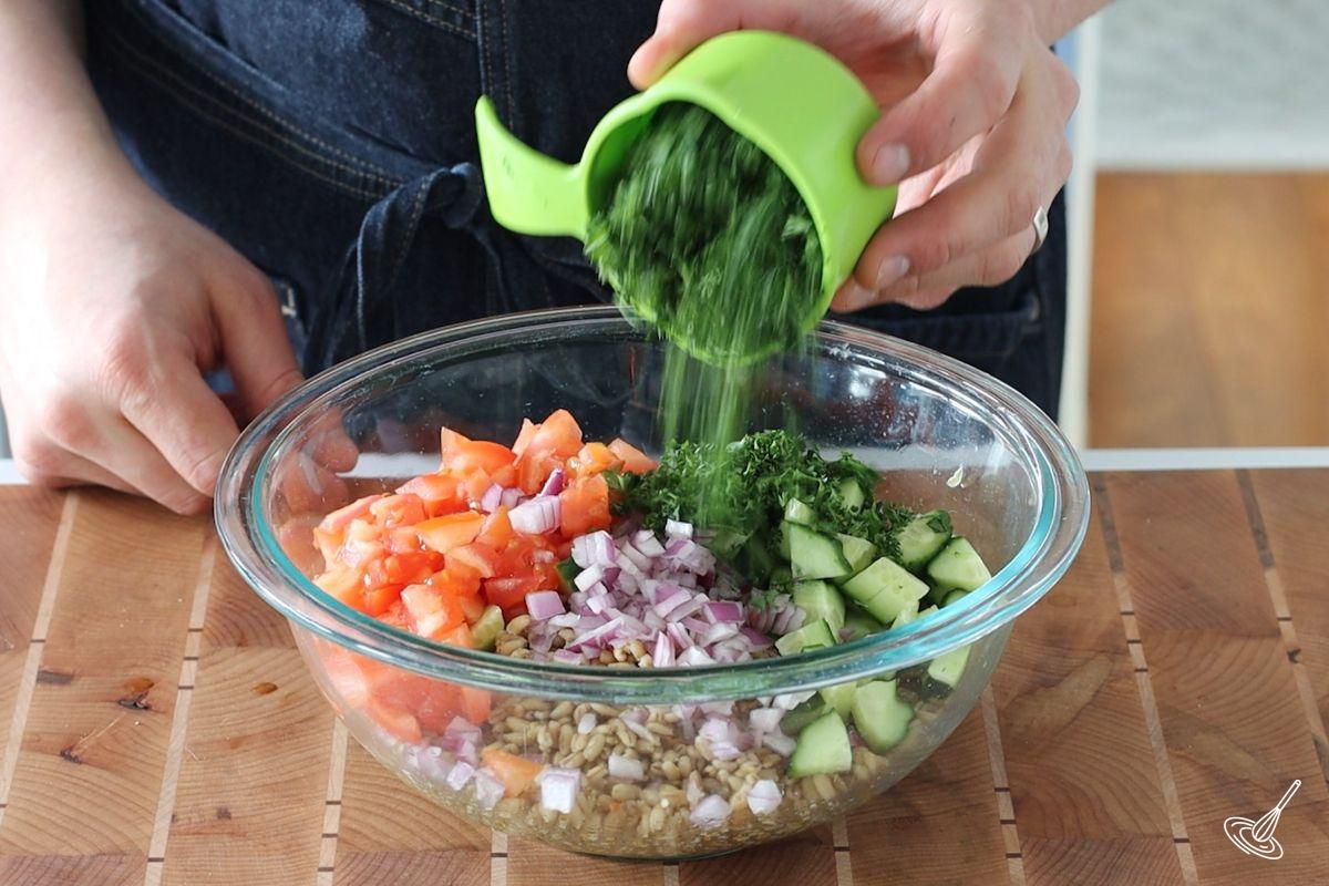 Someone adding chopped parsley to a large bowl of barley tabbouleh salad.
