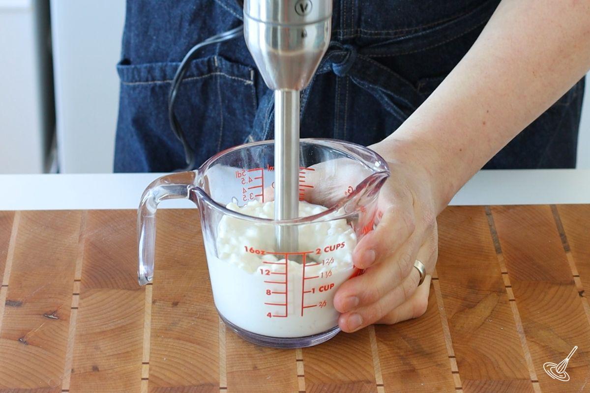 Someone using a stick blender to blend cottage cheese.