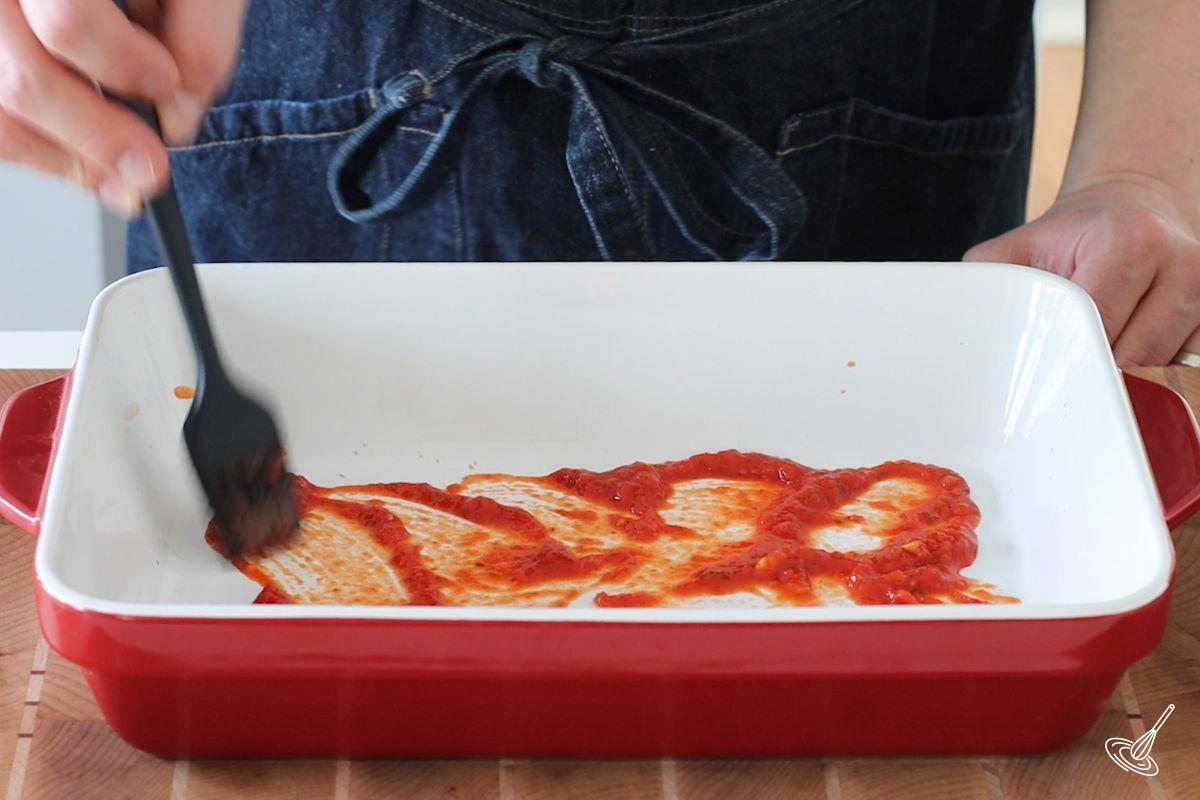 Someone brushing tomato sauce on the bottom of a 9x13 baking dish.