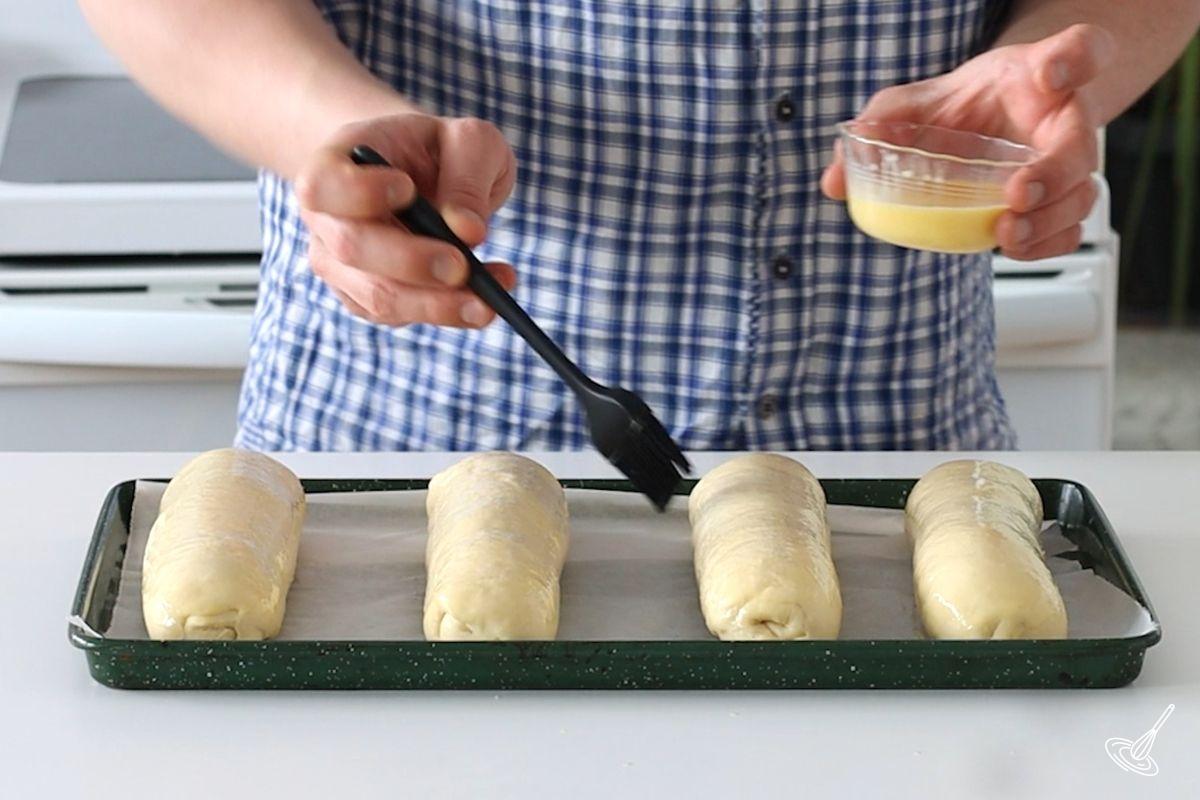 Someone brushing egg wash on brioche sub rolls.