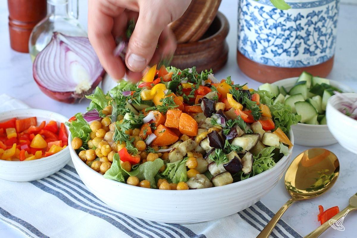 Someone adding fresh veggies on top of a Roasted Sweet Potato and Aubergine Salad with Tahini Dressing.