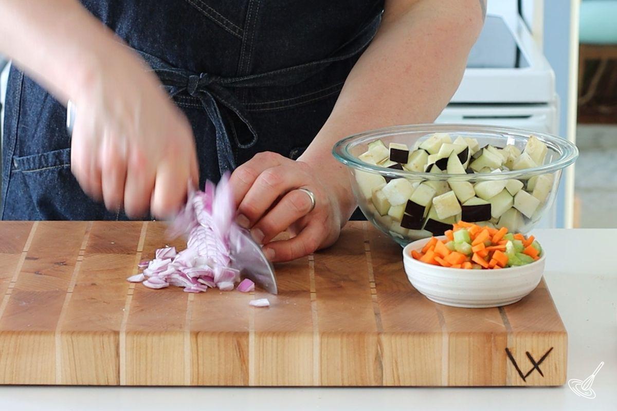 Someone using a knife to chop up red onion on a wooden cutting board.