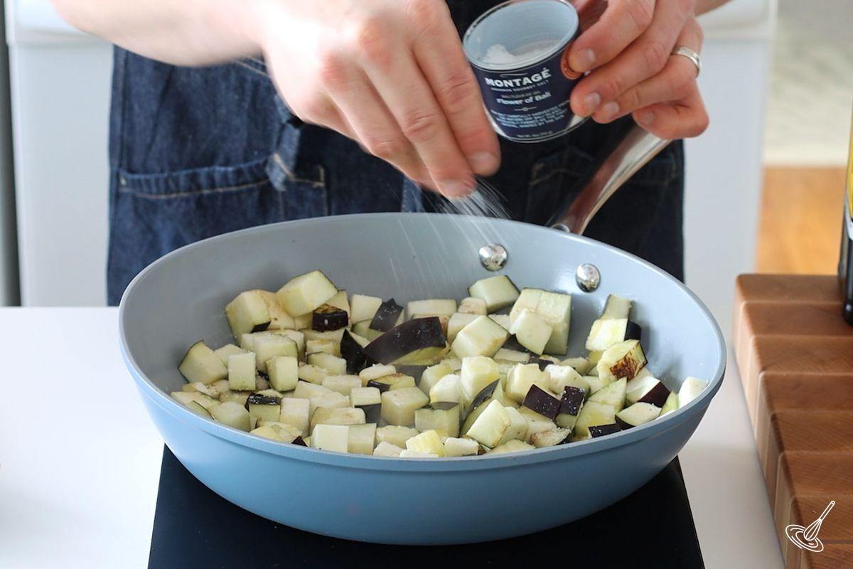 Someone sprinkling salt over cooking cubes of aubergines in a large pan. 