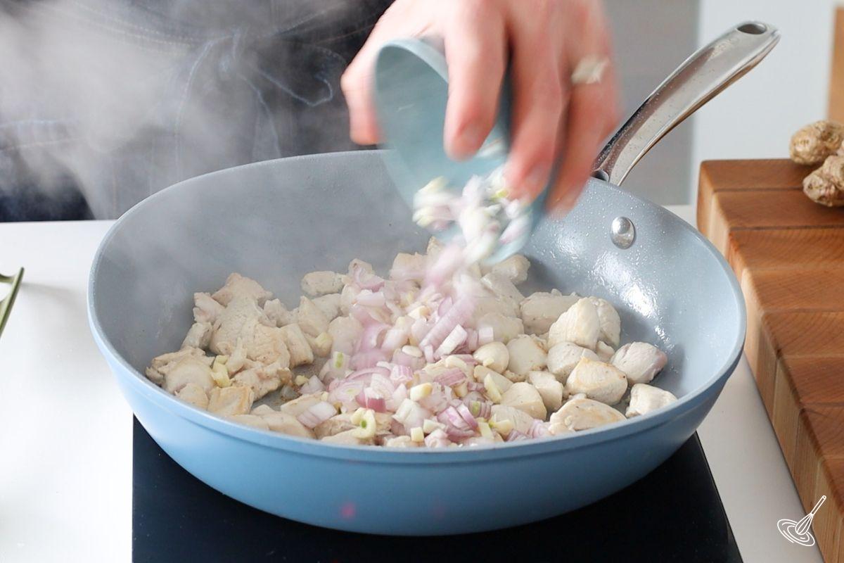 Someone adding garlic to a large pan of cooked chicken cubes. 