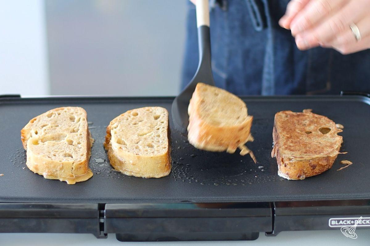 Someone using a spatula to flip over a cooked French toast on a griddle.