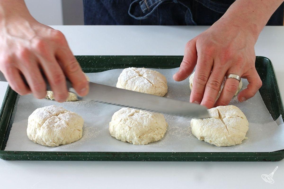 Someone using a bread knife to cut a cross on top of Irish soda bread rolls. 