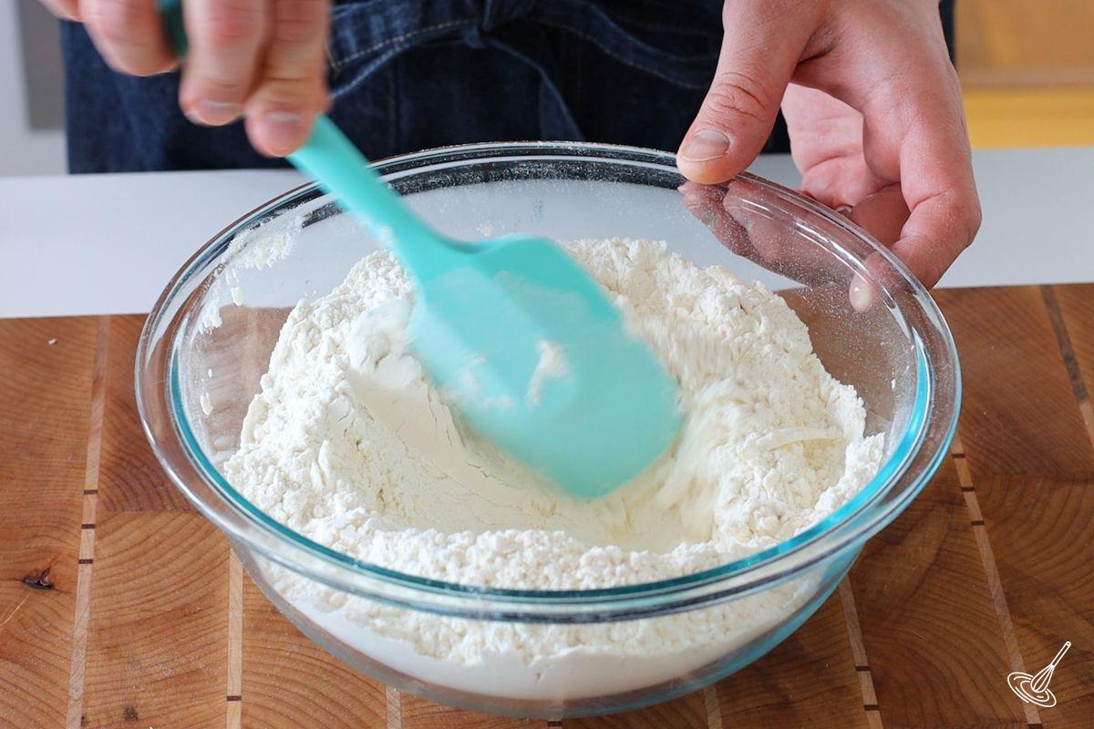 Someone using a rubber spatula to make a well in the center of a large bowl of flour. 