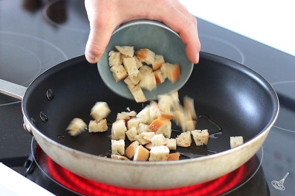 Someone adding cubes of bread to a large frying pan with olive oil.