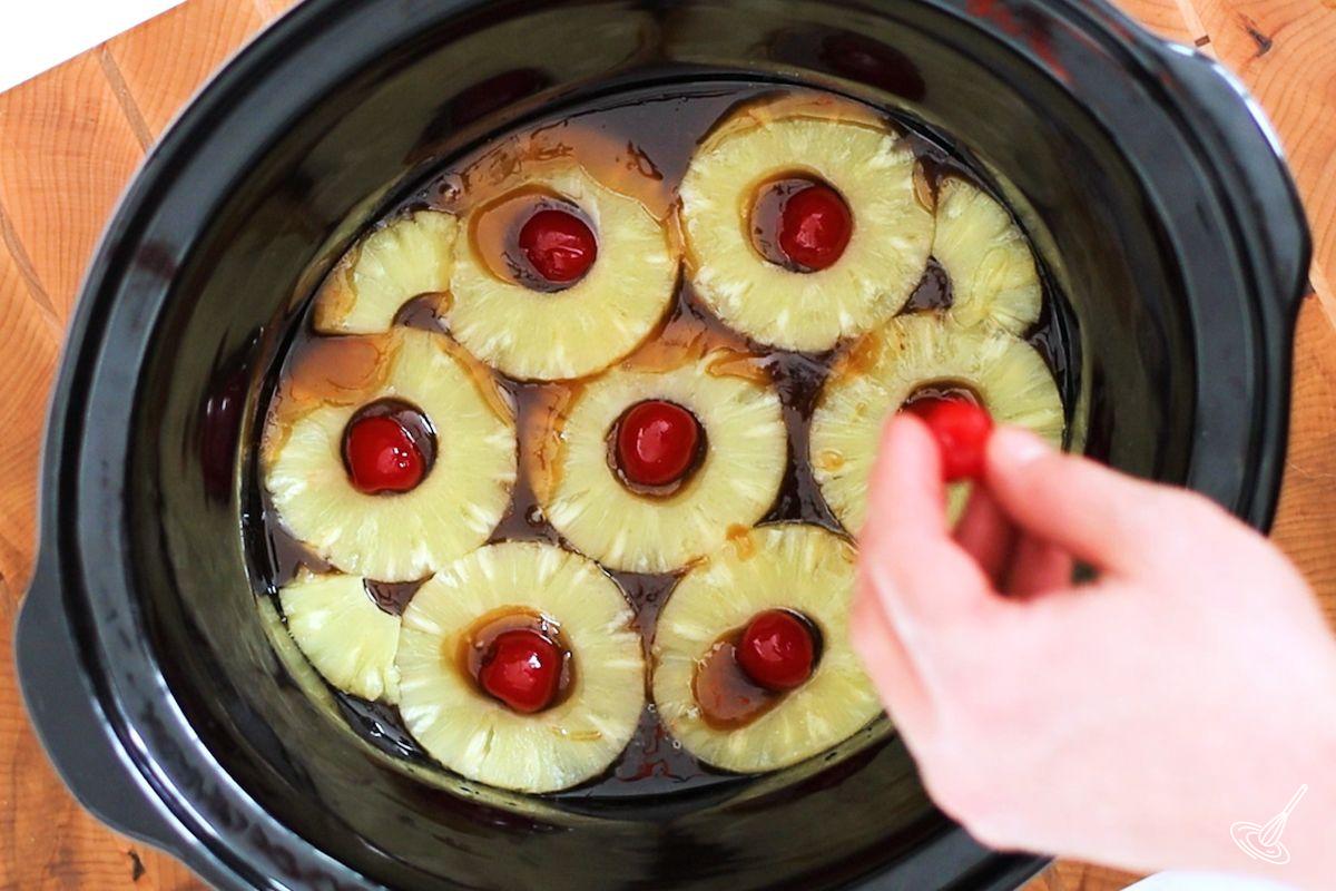 Someone placing pineapple slices and maraschino cherries on top of caramel in slow cooker crock.