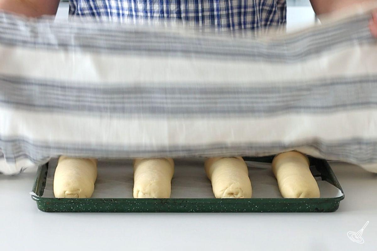 Someone placing a linen cloth over a tray of brioche sub roll recipe on a baking tray. 