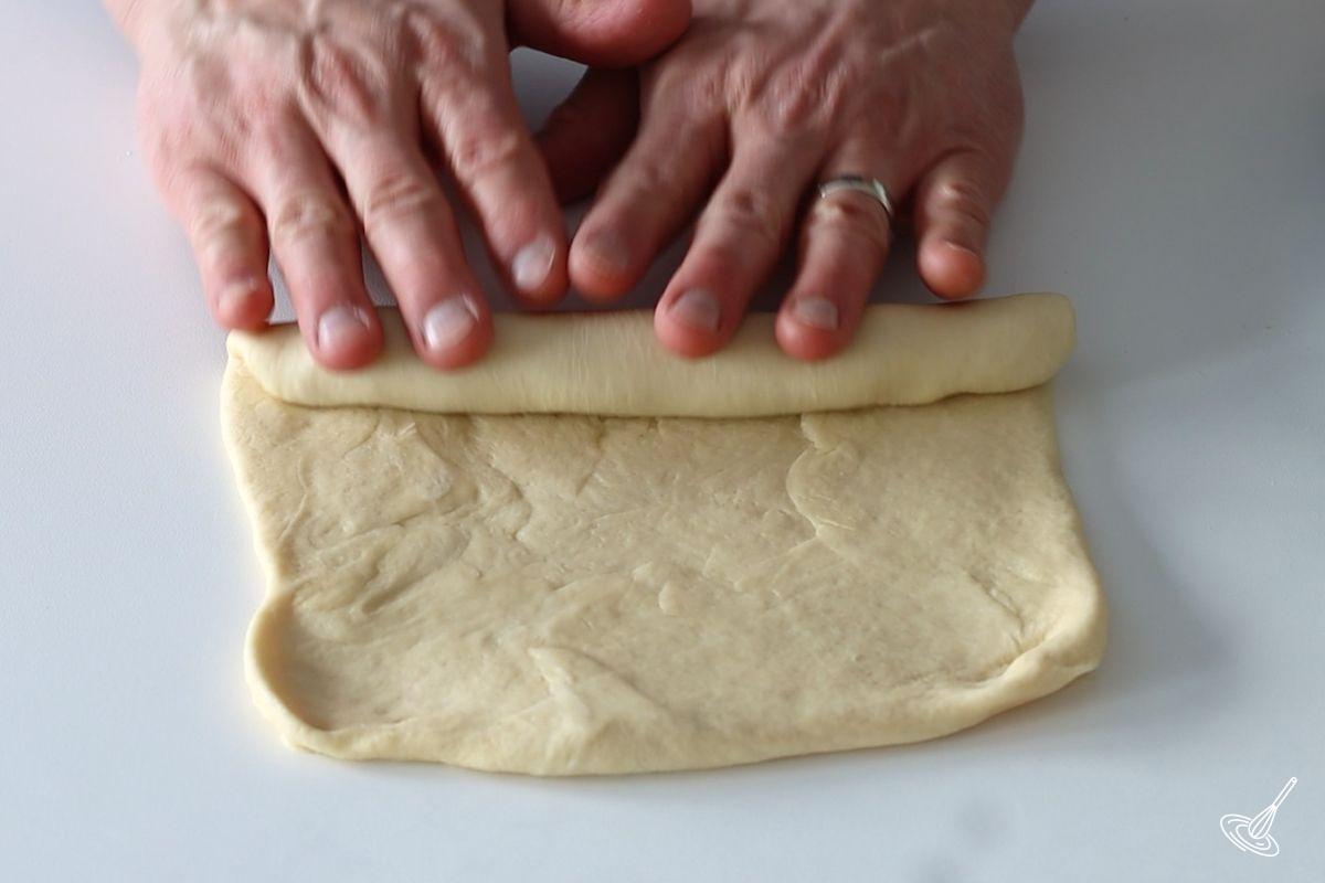 Someone using their two hands to roll up a flat square of dough. 