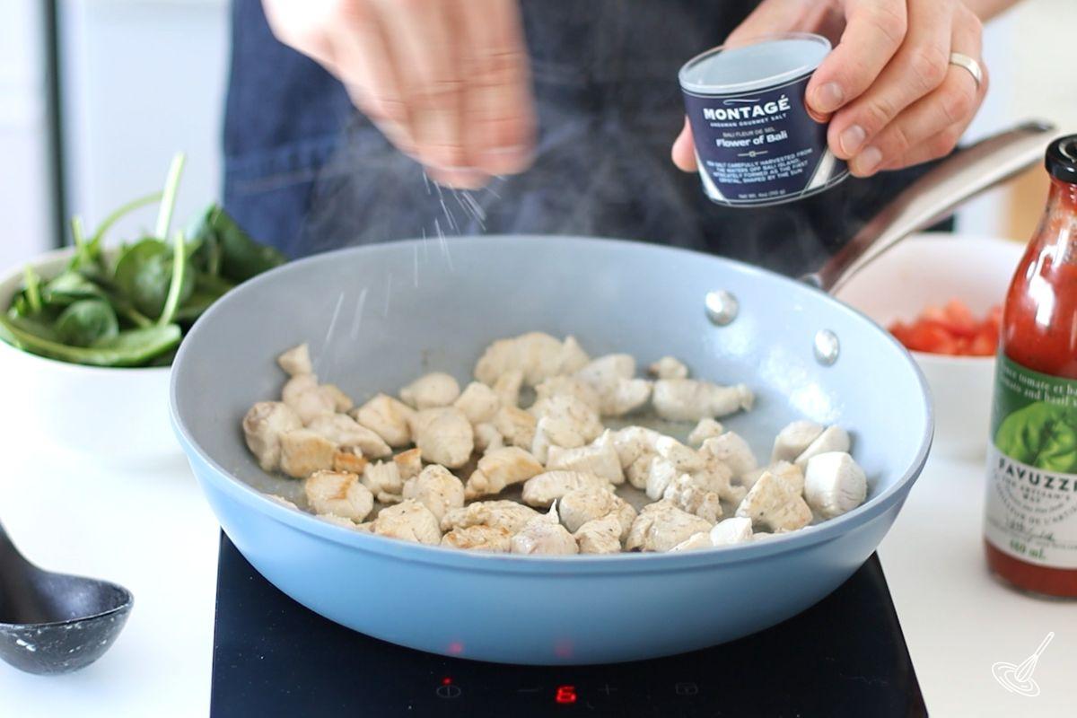 Someone sprinkling salt on cooked chicken cubes in a large skillet.