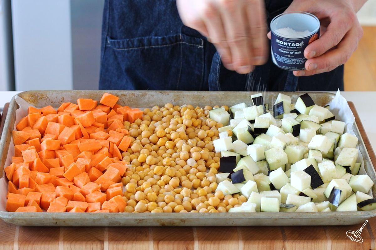 Someone sprinkling salt over a baking tray of cubes veggies. 