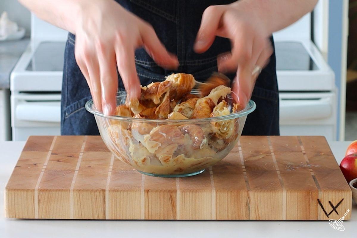 Someone using their two hands to toss pieces of croissant in a bowl of egg mixture.