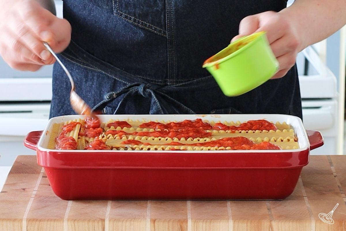 Someone spooning tomato sauce on top of lasagna noodles in a baking dish. 