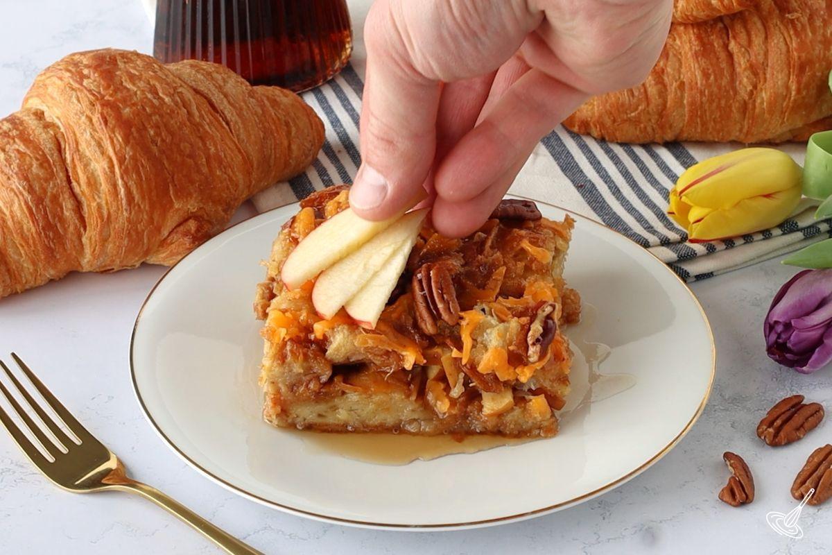 Someone placing sliced apple on top of a piece of Maple Croissant Bake.
