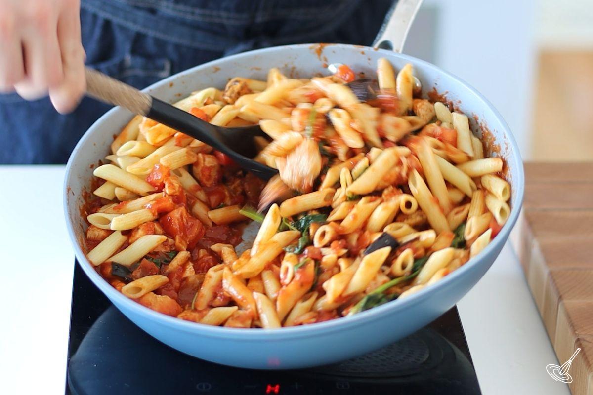 Someone stirring cooked pasta into a skillet of aubergine and chicken sauce.