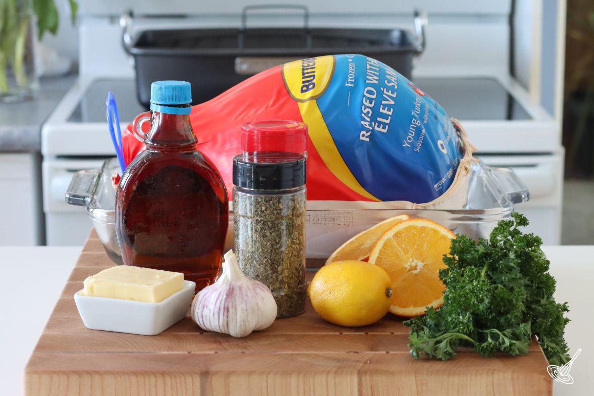Ingredients on the kitchen counter to make a roast turkey, including a whole turkey, maple syrup, herbs, garlic, orange, and butter.