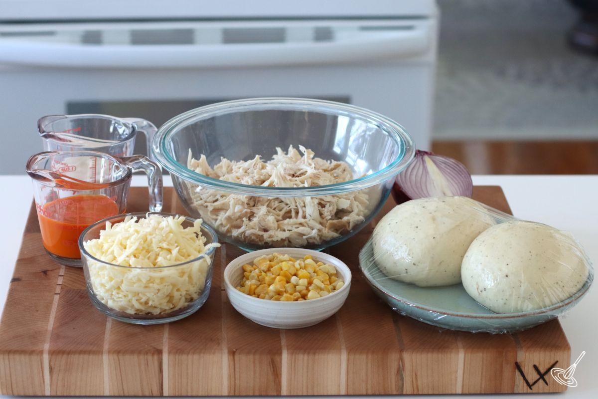 Ingredients on a cutting board including shredded turkey meat, pizza dough, grated cheese, and buffalo sauce. 
