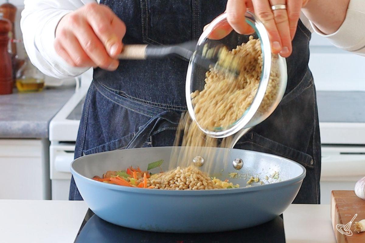 Someone adding cooked barley to a large pan of cooked vegetables.