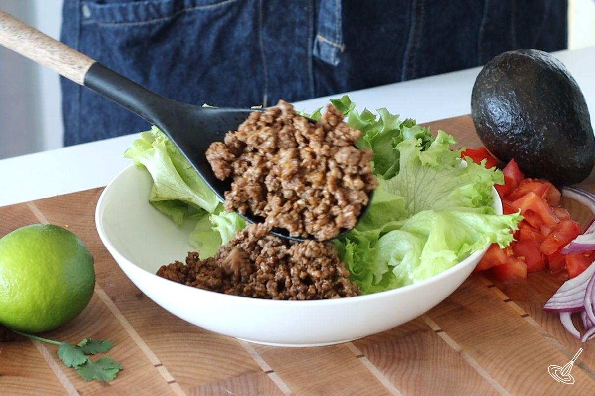 Someone adding a scoop of cooked taco beef to a serving bowl with lettuce. 