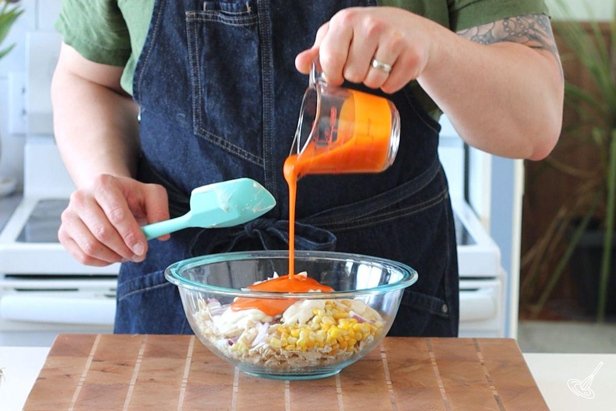 Someone adding buffalo sauce to a bowl of shredded turkey and veggies.