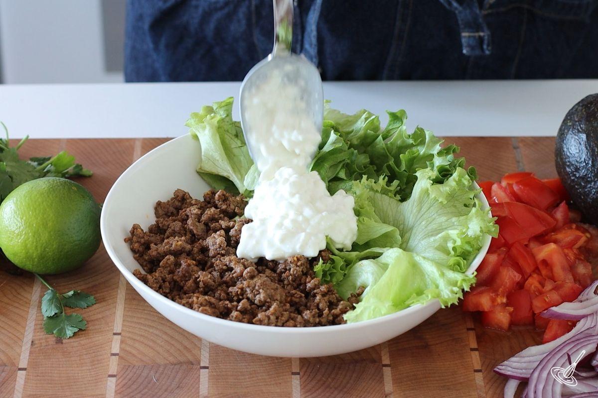 Someone adding a scoop of cottage cheese in a bowl with ground beef and lettuce. 