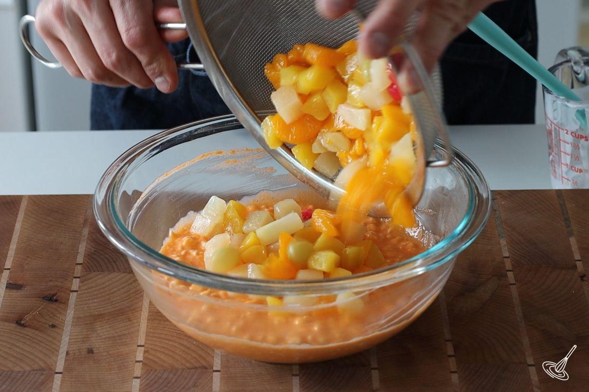 Someone adding drained fruit cocktail to a bowl of orange flavored cottage cheese.