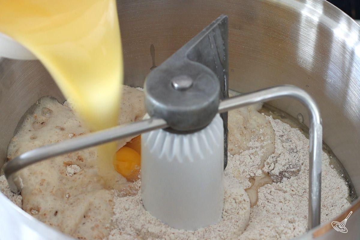 Someone pouring melted butter into the bowl of a stand mixer containing flour and eggs. 