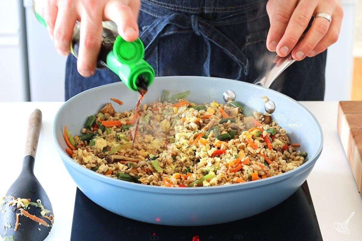 Someone pouring soy sauce in a large pan of barley fried rice. 