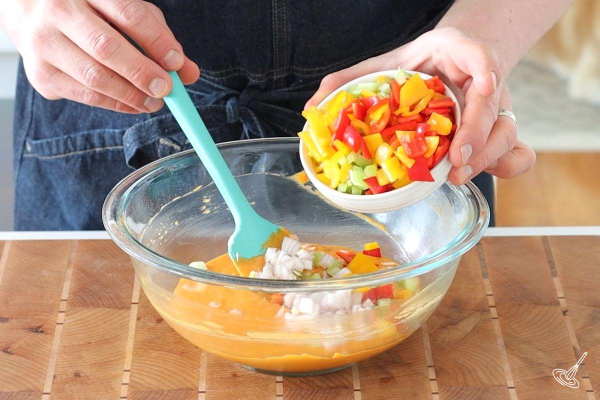 Someone adding a bowl of chopped vegetables to a large bowl of cheesy salad dressing.