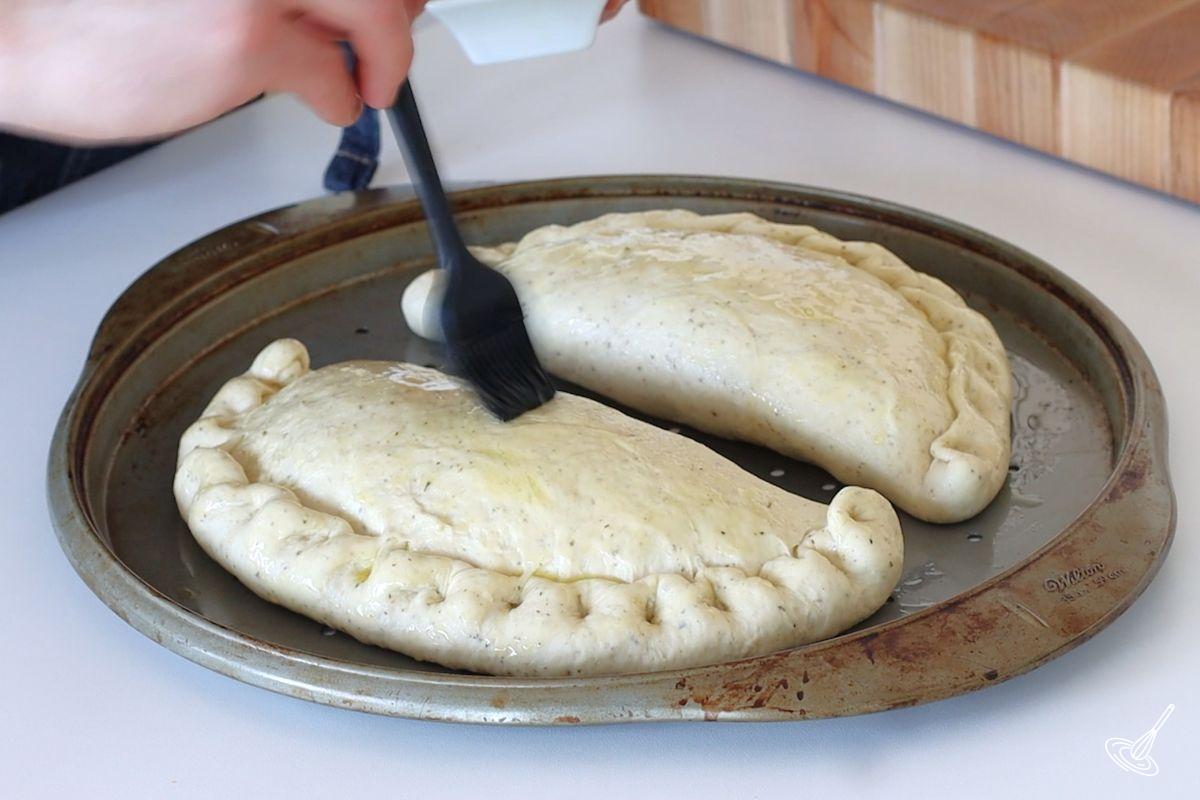 Someone brushing olive oil on shaped calzone before baking.