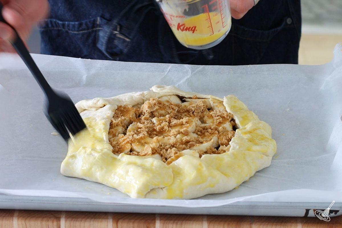 Someone brushing the edges of banana chocolate galette with egg wash.