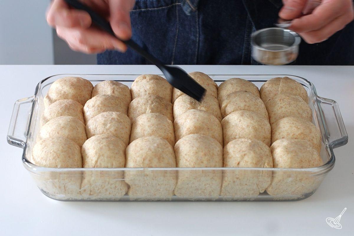 Someone using a silicone brush to brush water on top of proofed honey whole wheat dinner rolls.