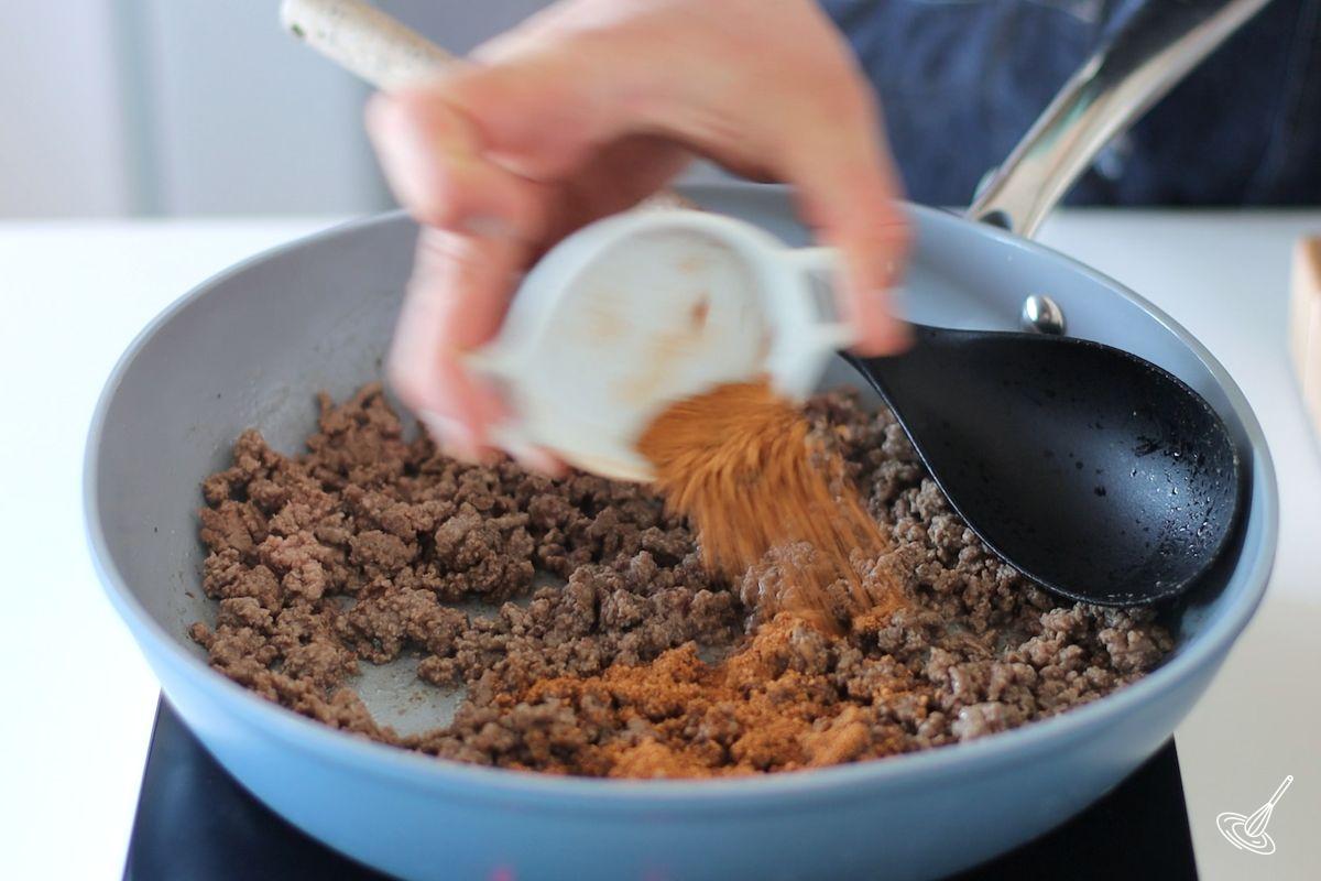 Someone adding taco seasoning to a frying pan of cooked ground beef.